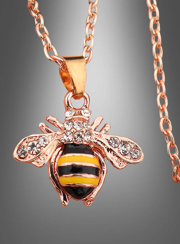 Entdecken Sie den „Bienen Kette“-Modeschmuck, eine goldene Halskette mit einem Bienenanhänger, der mit gelben, schwarzen und weißen Streifen sowie funkelnden Strasssteinen auf den Flügeln verziert ist. Vor einem grau abgestuften Hintergrund ist dieses bezaubernde Accessoire das perfekte Schmuckstück, um jedem Outfit eine verspielte Note zu verleihen.
