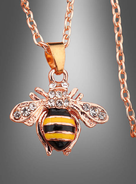 Entdecken Sie den „Bienen Kette“-Modeschmuck, eine goldene Halskette mit einem Bienenanhänger, der mit gelben, schwarzen und weißen Streifen sowie funkelnden Strasssteinen auf den Flügeln verziert ist. Vor einem grau abgestuften Hintergrund ist dieses bezaubernde Accessoire das perfekte Schmuckstück, um jedem Outfit eine verspielte Note zu verleihen.