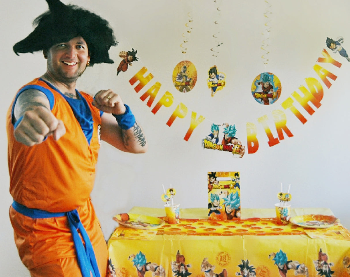 Eine Person in einem orangefarbenen Kostüm und einer schwarzen Stachelperücke posiert auf einer Geburtstagsparty zum Thema „Dragon Ball“, umgeben von farbenfrohen Tischdekorationen und den über den Köpfen hängenden „Dragon Ball Deko Swirls“ (5 Stück), wobei ein „Happy Birthday“-Banner die festliche Atmosphäre der Anime-Kindergeburtstagsparty abrundet.