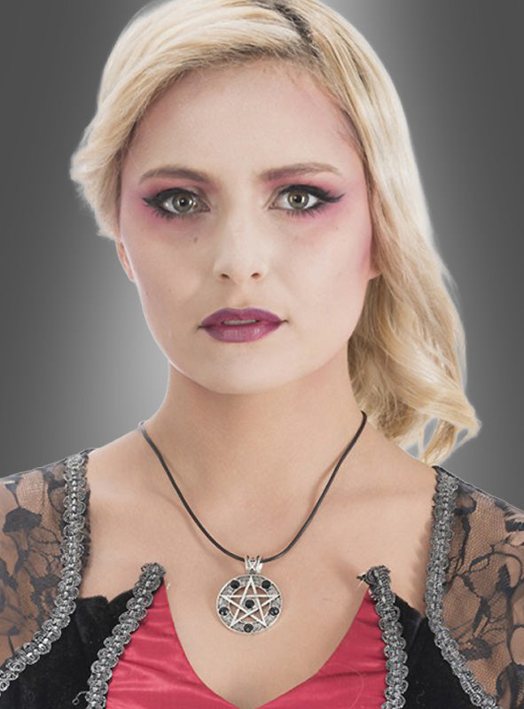 Eine Frau mit blonden Haaren, auffälligem rosa Augen-Make-up und schwarz-roter Kleidung mit Spitzenbesatz posiert vor einem grauen Hintergrund mit Farbverlauf und präsentiert dabei den markanten gotischen Schmuck der „Pentagramm-Amulett-Kette mit schwarzen Steinen“.