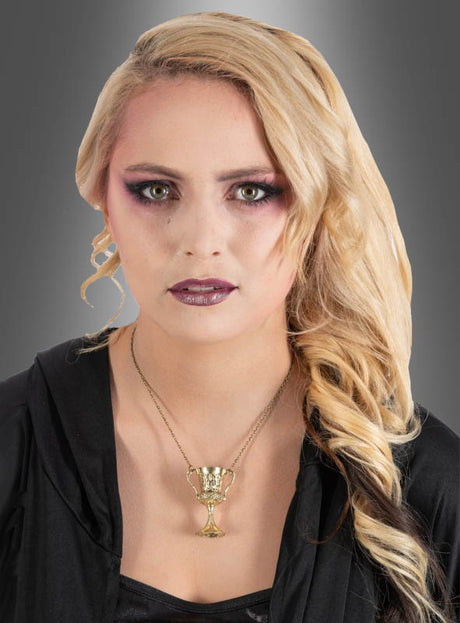 Eine Frau mit langen blonden Haaren, violettem Augen-Make-up und dunklem Lippenstift trägt ein schwarzes Oberteil und die „Goldene Kette mit Kelch-Anhänger“ – deren Kelch-Anhänger an die Magie von Harry Potter erinnert –, während sie vor einem grauen Hintergrund mit Farbverlauf posiert.