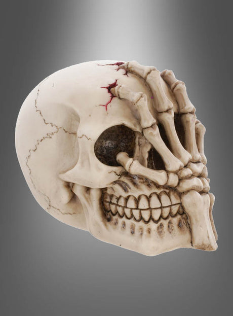 Das „Totenkopf mit Hand 12 x 15 cm Deluxe“-Modell zeigt einen menschlichen Schädel, dessen Mund und Kiefer von einer skelettierten Hand aus Knochen bedeckt sind. Ein markanter, rot gefärbter Riss im oberen Bereich setzt einen besonderen Akzent – ideal für Ihre nächste Halloween-Dekoration mit Totenköpfen.