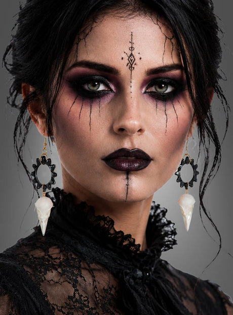 Eine Frau mit auffälligem Make-up um Augen und Lippen, die ein Spitzenoberteil trägt und deren schwarzes Haar hochgesteckt ist, blickt direkt in die Kamera. Abgerundet wird ihr Look durch „Skelett Ohrringe Krähe“ – Ohrringe in Form eines Krähen-Schädels.