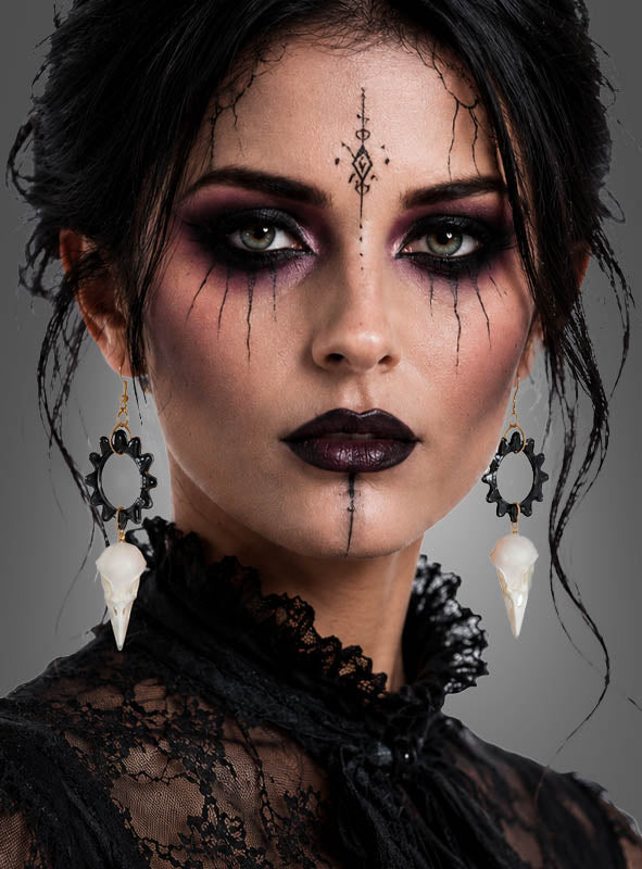 Eine Frau mit auffälligem Make-up um Augen und Lippen, die ein Spitzenoberteil trägt und deren schwarzes Haar hochgesteckt ist, blickt direkt in die Kamera. Abgerundet wird ihr Look durch „Skelett Ohrringe Krähe“ – Ohrringe in Form eines Krähen-Schädels.