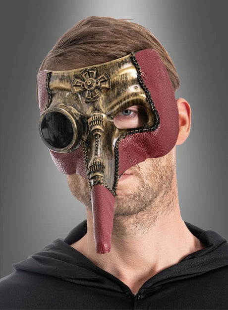 Ein Mann, der die „Steampunk Pestdoktor“-Maske trägt – eine Halbmaske mit bronzefarbenen Metallic- und roten Akzenten, die sich durch eine lange Nase und runde Augenöffnungen auszeichnet –, steht vor einem grauen Hintergrund.