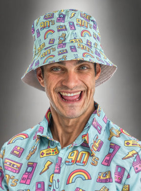 Ein lächelnder Mann trägt ein farblich passendes, farbenfrohes Shirt im 90er-Jahre-Stil und einen blauen Eimerhut mit 90er-Jahre-Motiv, der auf einem grauen Farbverlaufshintergrund mit Comicfiguren, Regenbögen und Kassetten verziert ist. Dieses lustige Retro-Party-Accessoire rundet seinen nostalgischen Look perfekt ab.