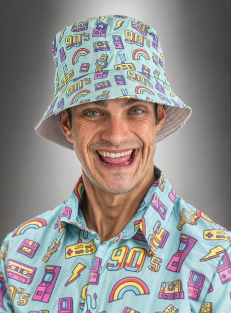 Ein lächelnder Mann trägt ein farblich passendes, farbenfrohes Shirt im 90er-Jahre-Stil und einen blauen Eimerhut mit 90er-Jahre-Motiv, der auf einem grauen Farbverlaufshintergrund mit Comicfiguren, Regenbögen und Kassetten verziert ist. Dieses lustige Retro-Party-Accessoire rundet seinen nostalgischen Look perfekt ab.