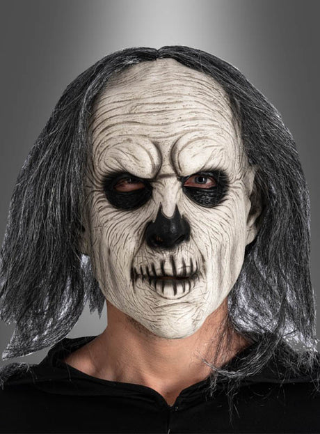 Eine Person, die die „Gruseliger Mann“-Latexmaske mit Haaren trägt – eine furchterregende Skelettmaske mit langen grauen Haaren, schwarzen Augen und Zahnabdrücken –, steht vor einem grauen Hintergrund mit Farbverlauf.