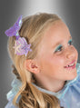 Ein junges Mädchen mit blonden Haaren trägt eine „Wunderschöne Meerjungfrau“-Haarspange und hellblaues Make-up; sie lächelt und blickt vor einem grauen Hintergrund mit Farbverlauf zur Seite.