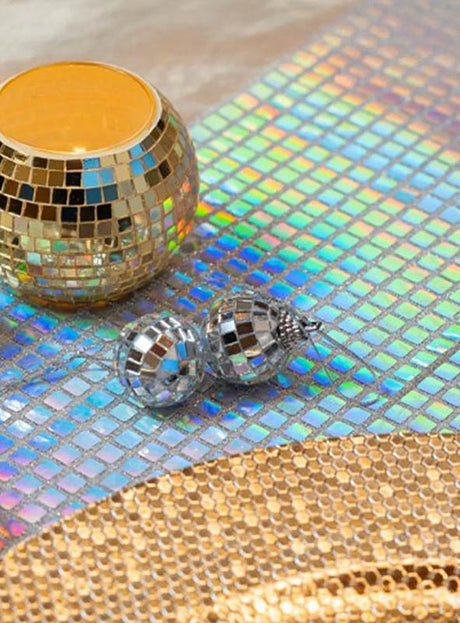 Gold- und silberfarbene Discokugel-Anhänger ruhen auf einer reflektierenden, schillernden Oberfläche mit Gittermuster, im Vordergrund befindet sich eine goldene Platte – ein glamouröses Discokugel-Design, das perfekt zum „Schillernder Disco Tischläufer silber 26x300cm“ passt.