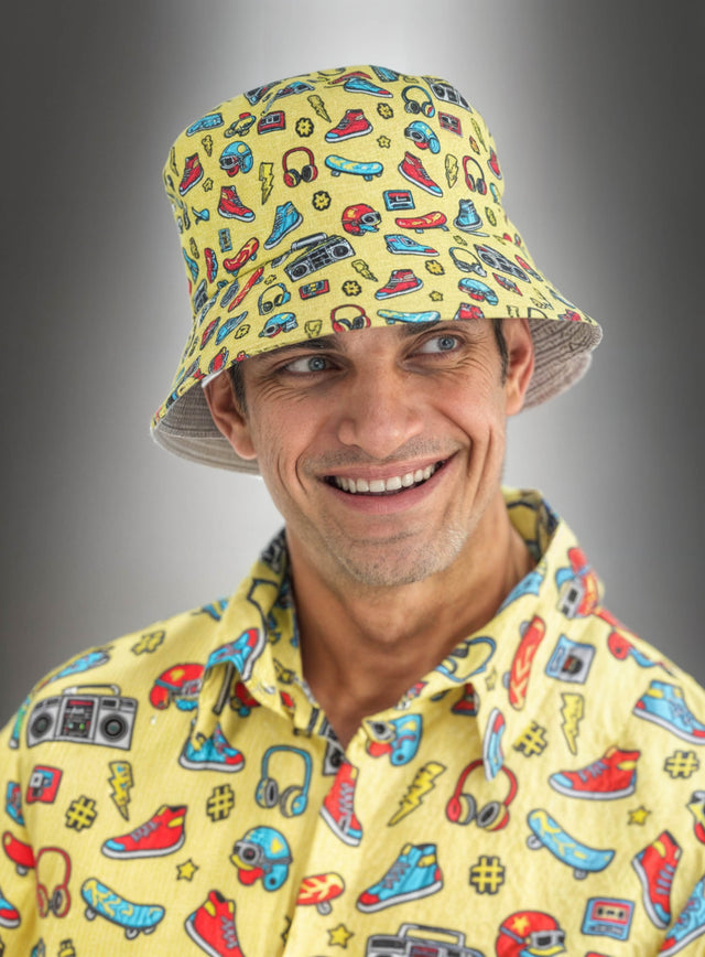 Ein lächelnder Mann trägt den gelben Eimerhut mit 90er-Jahre-Motiv, der perfekt zu seinem T-Shirt passt, das mit bunten Comic-Motiven wie Schuhen, Ghettoboxen, Kopfhörern und Sonnenbrillen bedruckt ist – ein echtes Retro-Party-Accessoire mit lebhaften 90er-Motiven.