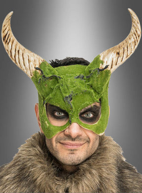 Eine Person, die die „Gehörnte Maske mit Moos-Effekt“ trägt – ein grünes, moosartiges Design mit Hörnern – sowie Pelzkleidung, blickt mit dunklem Augen-Make-up vor einem grauen Hintergrund mit Farbverlauf in die Kamera.