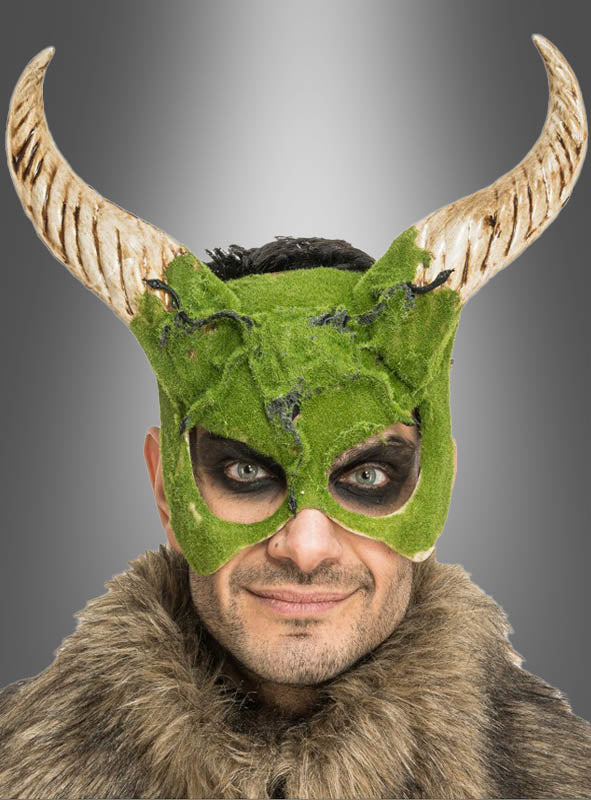 Eine Person, die die „Gehörnte Maske mit Moos-Effekt“ trägt – ein grünes, moosartiges Design mit Hörnern – sowie Pelzkleidung, blickt mit dunklem Augen-Make-up vor einem grauen Hintergrund mit Farbverlauf in die Kamera.