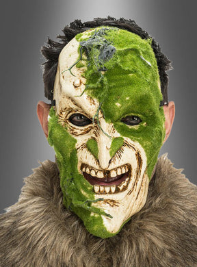 Eine Person, die die „Gruselige Maske mit Moos-Effekt“ trägt – eine zur Hälfte aus grünem Moos bestehende, zur Hälfte gruselige Horrormaske mit übertriebenen Gesichtszügen –, kombiniert mit einem Kleidungsstück mit Pelzkragen, blickt vor einem grauen Hintergrund in die Kamera.
