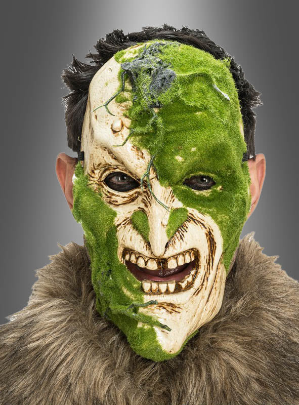 Eine Person, die die „Gruselige Maske mit Moos-Effekt“ trägt – eine zur Hälfte aus grünem Moos bestehende, zur Hälfte gruselige Horrormaske mit übertriebenen Gesichtszügen –, kombiniert mit einem Kleidungsstück mit Pelzkragen, blickt vor einem grauen Hintergrund in die Kamera.