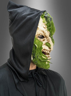 Eine Person in einem schwarzen Kapuzenmantel, die die „Gruselige Maske mit Moos-Effekt“ trägt – eine grüne und beigefarbene, monströse Maske –, im Seitenprofil vor grauem Hintergrund.