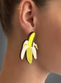 Nahaufnahme des Ohrs einer Person, die große, farbenfrohe Acryl-Ohrringe in Form einer Banane sowie einen kleinen goldenen Creolenring im oberen Ohrbereich trägt, was den verspielten Modeschmuck-Stil unterstreicht.