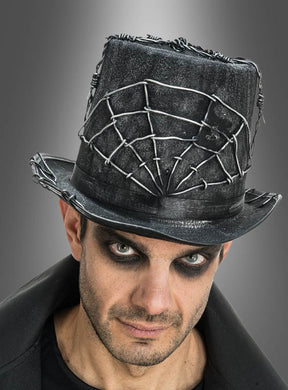 Ein Mann mit auffälligem Augen-Make-up trägt den „Halloween-Zylinder mit Draht-Spinnennetz“ – einen schwarzen Hut, der mit Stacheldraht und Spinnennetz-Motiven verziert ist und vor einem schlichten grauen Hintergrund zur Geltung kommt – ein auffälliges Kostüm-Accessoire, das sich ideal für gruselige Anlässe eignet.