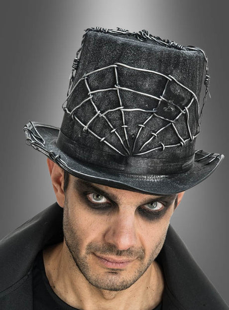 Ein Mann mit auffälligem Augen-Make-up trägt den „Halloween-Zylinder mit Draht-Spinnennetz“ – einen schwarzen Hut, der mit Stacheldraht und Spinnennetz-Motiven verziert ist und vor einem schlichten grauen Hintergrund zur Geltung kommt – ein auffälliges Kostüm-Accessoire, das sich ideal für gruselige Anlässe eignet.