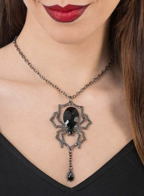 Eine Frau in einem schwarzen Oberteil und mit dunklem Lippenstift präsentiert die „Schwarze Spinne“-Halskette mit Edelstein, deren Blickfang eine markante schwarze Edelstein-Spinne mit einem herabhängenden, tropfenförmigen Edelstein ist – damit ist sie der perfekte Halloween-Schmuck.