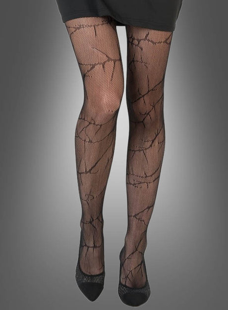 Eine Person, die eine schwarze gemusterte Strumpfhose – genauer gesagt die „Strumpfhose Dornen Ranken Muster“ – und einen schwarzen Rock trägt und vor einem grauen Hintergrund steht. Zu sehen sind nur der Unterkörper und die Füße in schwarzen Schuhen.