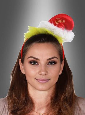 Eine Frau mit braunen Haaren trägt die Original-Grinch-Haarreif-Mütze, ein rot-weißes Stirnband mit der Aufschrift „The Grinch“ und grünem Fell, und posiert vor einem grau verlaufenden Hintergrund.