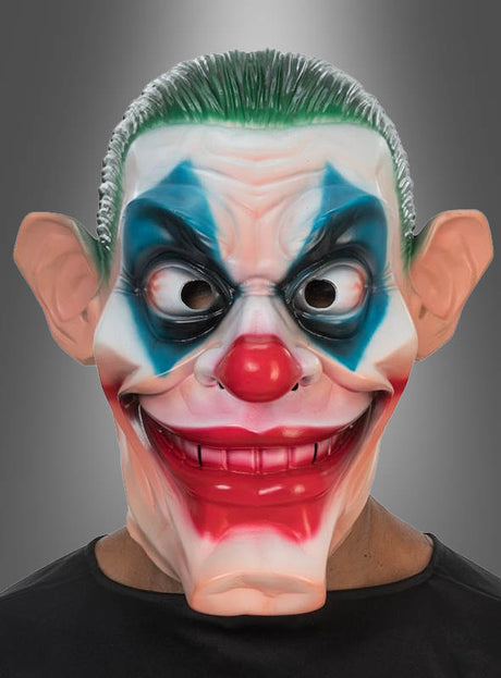 Eine Person, die die „Horror Clown Joker“-Vollmaske trägt – mit übertrieben großen Ohren, grünen Haaren, blauem Augen-Make-up, einer roten Nase und einem breiten, aufgemalten Grinsen –, steht vor einem grauen Hintergrund.