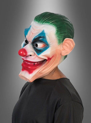 Eine Person, die die Horror-Clown-Joker-Vollmaske trägt, mit leuchtend grünem Haar, einer roten Nase, auffälligem blauem Augen-Make-up und einem übertriebenen roten Lächeln. Die Schaumlatex-Konstruktion dieser Schaumstoffmaske sorgt für ein auffallend realistisches Joker-Erscheinungsbild. Der Hintergrund ist ein schlichter grauer Farbverlauf.
