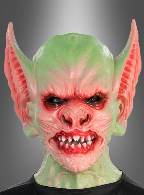 Eine Person, die die „Goblin Kobold Schaumstoff-Vollmaske Deluxe“ trägt – mit einem grün-roten Design, großen spitzen Ohren, faltiger Haut, scharfen Zähnen und einem wütenden Gesichtsausdruck –, steht vor einem grauen Hintergrund mit Farbverlauf.