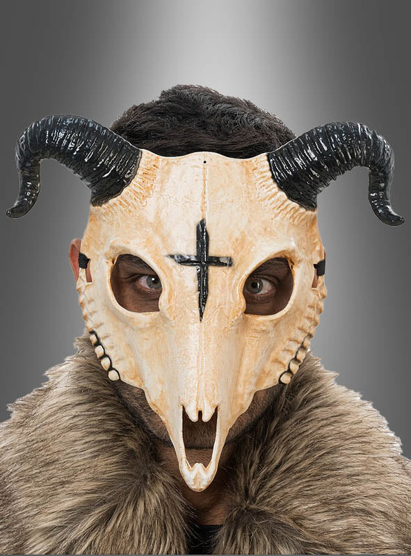Eine Person, die die „Gehörnte Tierschädel-Halbmaske“ trägt – eine Halbmaske in Form eines gehörnten Tierschädels mit einem schwarzen Kreuz auf der Stirn –, gekleidet in ein Pelzgewand, vor einem grauen Hintergrund.