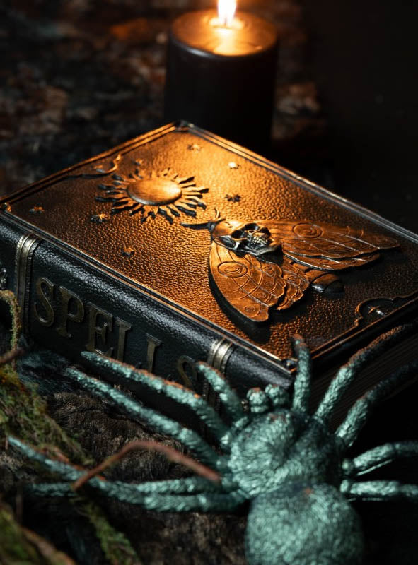 Die Zauberbuch-Dekoration „Grimoire“ (20 × 15 cm) – ein aus Leder gefertigtes „Buch der Schatten“ mit der Aufschrift „Spells“ und einem geprägten Motiv aus Motte und Sonne – ruht neben einer brennenden schwarzen Kerze und einer kunstvoll gestalteten Spinne und ist damit die ideale Zauberbuch-Dekoration für Halloween oder Hexenbuch-Dekoration für Ihr magisches Arrangement.