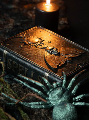 Die Zauberbuch-Dekoration „Grimoire“ (20 × 15 cm) – ein aus Leder gefertigtes „Buch der Schatten“ mit der Aufschrift „Spells“ und einem geprägten Motiv aus Motte und Sonne – ruht neben einer brennenden schwarzen Kerze und einer kunstvoll gestalteten Spinne und ist damit die ideale Zauberbuch-Dekoration für Halloween oder Hexenbuch-Dekoration für Ihr magisches Arrangement.
