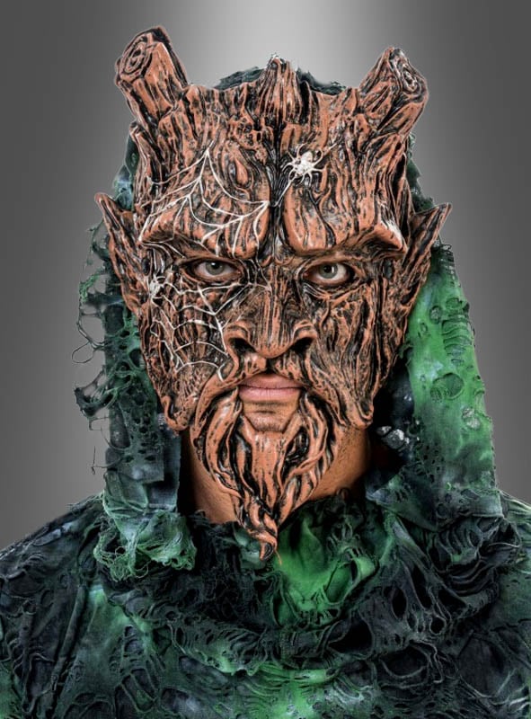Eine Person, die die „Waldgeist Baum“-Halbmaske trägt – die so gestaltet ist, dass sie wie Baumrinde mit Hörnern und Spinnweben aussieht – und dazu ein grünes, zerfleddertes Kostüm trägt, blickt vor einem schlichten grauen Hintergrund nach vorne.