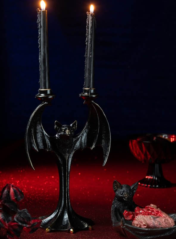 Ein „Fledermaus“-Kerzenständer (35 cm) aus Kunstharz in Form einer schwarzen Fledermaus, auf dem zwei brennende schwarze Kerzen stehen, ruht auf einer roten Unterlage neben einer kleinen Fledermausfigur und einer Schale mit Fleisch – ideal als Gothic-Deko oder als auffällige Halloween-Tischdekoration.