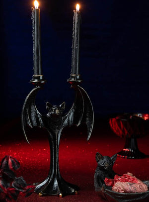 Ein „Fledermaus“-Kerzenständer (35 cm) aus Kunstharz in Form einer schwarzen Fledermaus, auf dem zwei brennende schwarze Kerzen stehen, ruht auf einer roten Unterlage neben einer kleinen Fledermausfigur und einer Schale mit Fleisch – ideal als Gothic-Deko oder als auffällige Halloween-Tischdekoration.
