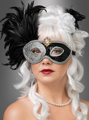 Eine Frau mit lockigem weißem Haar trägt die „Venezianische Maske schwarz silber“, die mit eleganten schwarzen und silbernen Federn verziert ist. Außerdem trägt sie eine weiße Blume im Haar, roten Lippenstift und eine Perlenkette – all dies vor einem grauen Hintergrund.