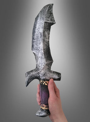 Eine Hand hält das „Schaumstoff Messer Dolch 39 cm“ – ein großes, verwittertes Schwert im Fantasy-Stil mit einer gezackten Klinge und einem violett-goldenen Griff –, das sich ideal als Halloween-Accessoire oder Kostümrequisite eignet und vor einem grauen Hintergrund mit Farbverlauf zu sehen ist.