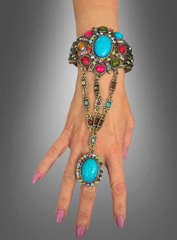 Eine Hand hält das „Orientalisches Armband aus Metall“ in den Händen, eine kunstvoll gestaltete Kombination aus Armband und Ring, die von orientalischem Armschmuck inspiriert ist. Sie ist mit bunten Edelsteinen und einem großen blauen Stein sowohl am Armband als auch am Ring verziert, die alle durch dekorative Goldketten elegant miteinander verbunden sind.