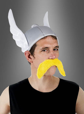 Eine Person, die den „Asterix-Helm – Original-Lizenz – Erwachsene“ trägt – inspiriert vom klassischen Flügelhelm-Design – sowie einen großen gelben Schnurrbart, gekleidet in ein schwarzes, ärmelloses Shirt und posierend vor einem grauen Hintergrund mit Farbverlauf – ein auffälliger Herrenkostüm-Look.