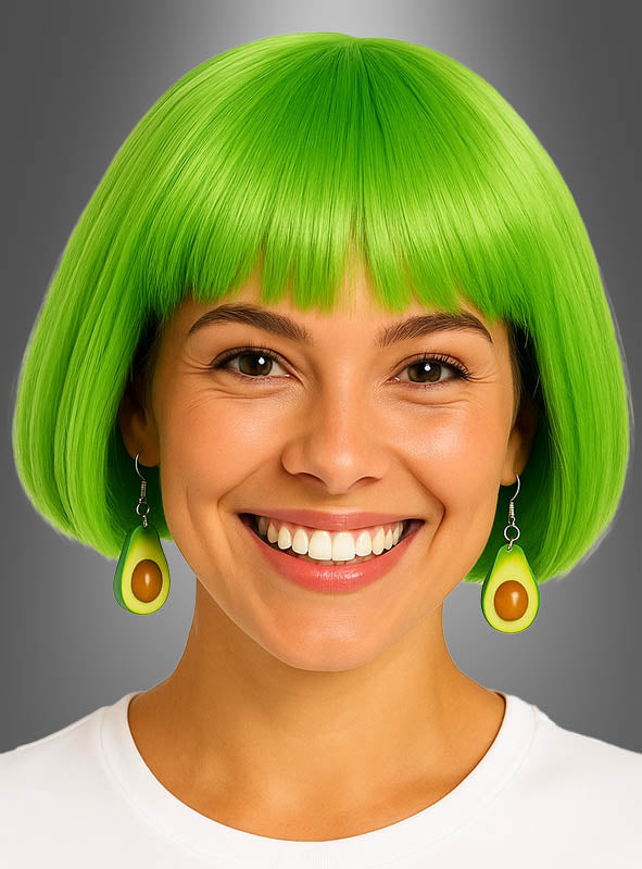 Eine lächelnde Person mit einem leuchtend grünen Bob-Haarschnitt, die Avocado-Ohrringe als Kostümschmuck und ein weißes Hemd trägt, vor einem grauen Hintergrund mit Farbverlauf.