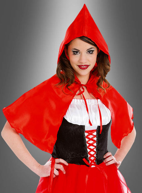 Eine Frau, die den „Roten Kurzumhang mit Kapuze Rotkäppchen“ – einen roten Umhang mit Kapuze – über einem schwarz-weißen Kleid mit roten Schnürungen trägt, steht mit den Händen in die Hüften gestemmt vor einem grauen Hintergrund mit Farbverlauf.