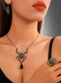 Eine Frau präsentiert das „Spinnen-Schmuckset mit grünem Edelstein“ – ein elegantes Accessoire im Gothic-Stil, bestehend aus passenden Ohrringen, einer Halskette und einem Ring mit Spinnenmotiv, die mit leuchtend grünen Edelsteinakzenten verziert sind – vor einem schlichten Hintergrund mit Farbverlauf.