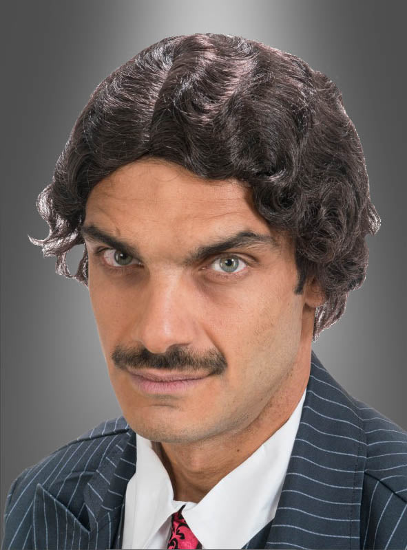Ein Mann mit welligem, dunklem Haar, das wie die Original-Gomez-Perücke aus „Wednesday“ frisiert ist, mit Schnurrbart und einem gestreiften Anzugjackett, das an die Addams Family erinnert, blickt vor einem grauen Hintergrund direkt in die Kamera.