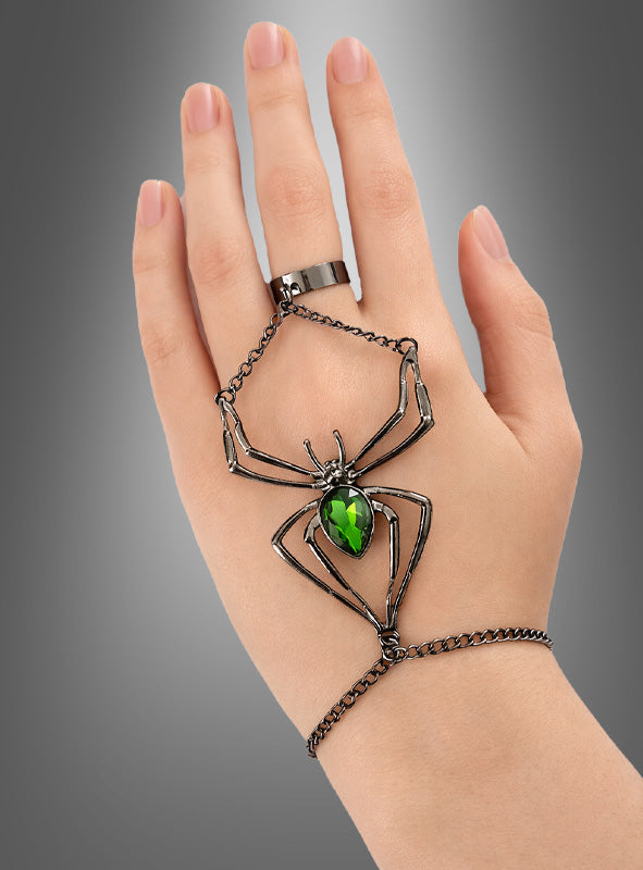 Eine Hand, geschmückt mit dem „Spinnen-Schmuck-Set mit grünem Edelstein“ – einem silbernen, spinnenförmigen Armbandring, bei dem ein großer grüner Edelstein den Körper der Spinne bildet –, glänzt als gotisches Accessoire auffallend vor einem grauen Hintergrund mit Farbverlauf.