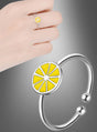 Das Set aus dem verstellbaren „Zitronen Ring“ und einem Armband mit gelbem Zitronenscheiben-Motiv: Der verstellbare „Zitronen Ring“ wird an einer Hand getragen und setzt einen verspielten Akzent als Modeschmuck, während das passende Armband separat vor einem grauen Farbverlaufshintergrund präsentiert wird.