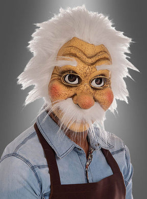 Eine Person, die die „Geppetto Opa Maske Deluxe“ aus „Pinocchio“ trägt – mit dem detailgetreuen Gesicht eines älteren Mannes mit weißem Haar und Schnurrbart –, ist in ein Jeanshemd und eine braune Schürze gekleidet und steht vor einem grauen Hintergrund.
