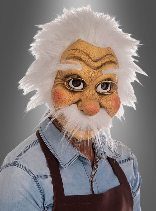 Eine Person, die die „Geppetto Opa Maske Deluxe“ aus „Pinocchio“ trägt – mit dem detailgetreuen Gesicht eines älteren Mannes mit weißem Haar und Schnurrbart –, ist in ein Jeanshemd und eine braune Schürze gekleidet und steht vor einem grauen Hintergrund.