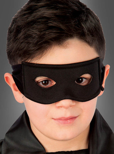Ein Kind mit kurzen, dunklen Haaren trägt die „Schwarze Augenmaske für Kinder u. Erwachsene“ und ein schwarzes Outfit und blickt vor einem schlichten grauen Hintergrund leicht zur Seite – und zeigt damit, wie dieses schlichte und doch auffällige Kostümaccessoire jeden Look aufwerten kann.