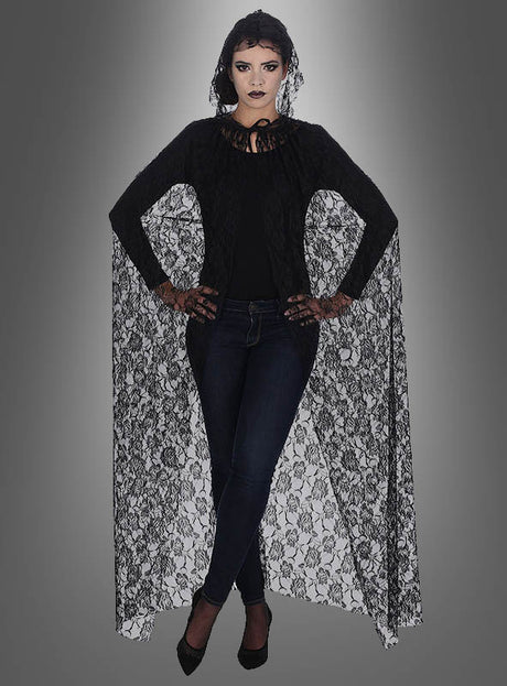 Eine Frau steht mit verschränkten Armen vor einem grau verlaufenden Hintergrund und trägt einen ärmellosen „Deluxe“-Umhang aus Spitze, ein schwarzes Oberteil, dunkle Jeans und schwarze Pumps. Ihr Make-up ist dramatisch und im Gothic-Stil gehalten – eine auffällige Wahl für ein Halloween-Kostüm.