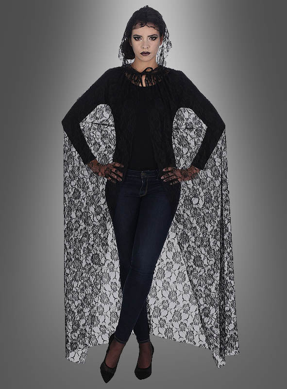 Eine Frau steht mit verschränkten Armen vor einem grau verlaufenden Hintergrund und trägt einen ärmellosen „Deluxe“-Umhang aus Spitze, ein schwarzes Oberteil, dunkle Jeans und schwarze Pumps. Ihr Make-up ist dramatisch und im Gothic-Stil gehalten – eine auffällige Wahl für ein Halloween-Kostüm.
